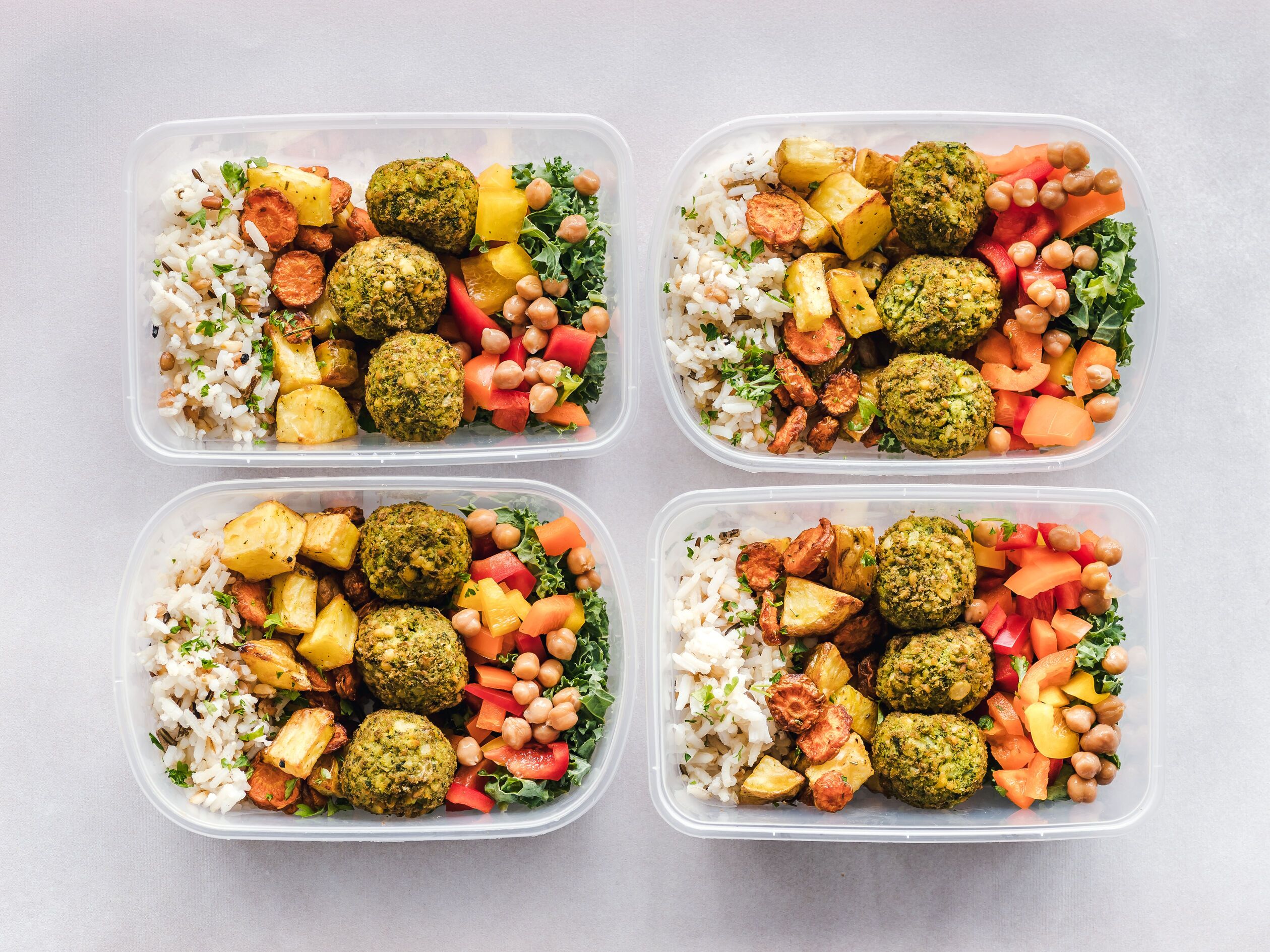 Four meal-prep containers with brown rice, roasted potatoes, chickpeas, peppers, greens and green falafel balls on a light grey table