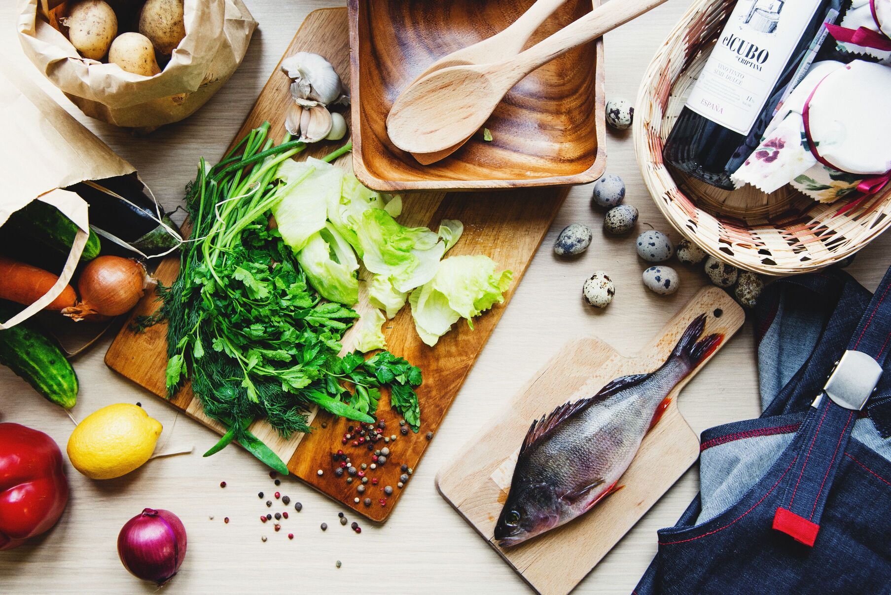 Fresh ingredients on a wooden board: leafy greens, lettuce, a whole fish, lemon, onion, garlic and wooden spoons