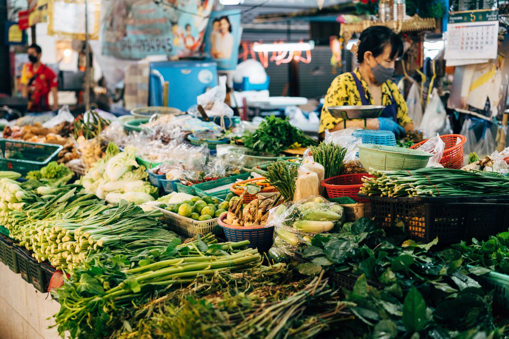 A busy Thai fresh market with heaps of green vegetables, herbs and a vendor wearing a mask