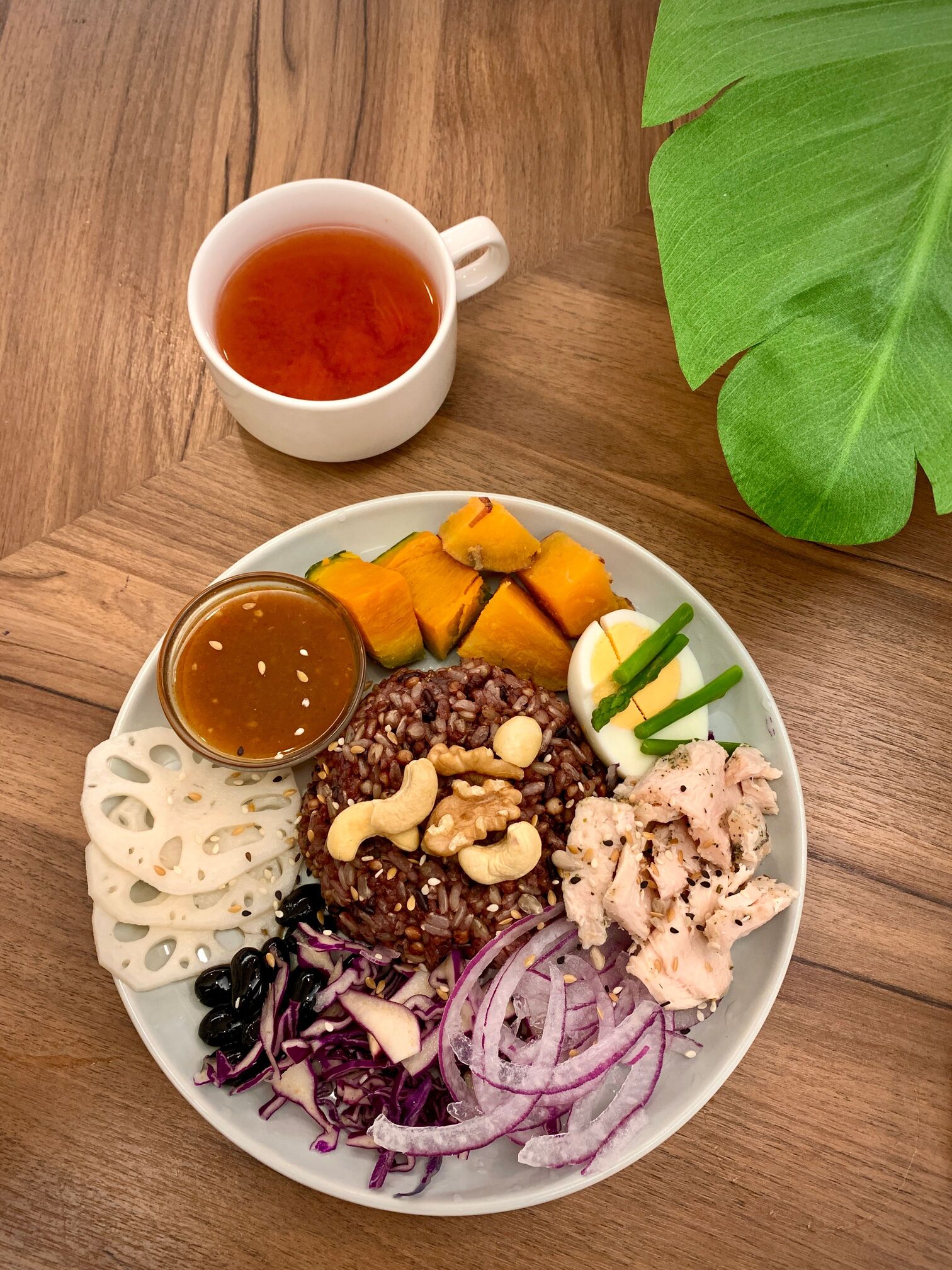 A balanced plate with riceberry rice, pumpkin, lotus root, egg, chicken and onions next to a cup of tea