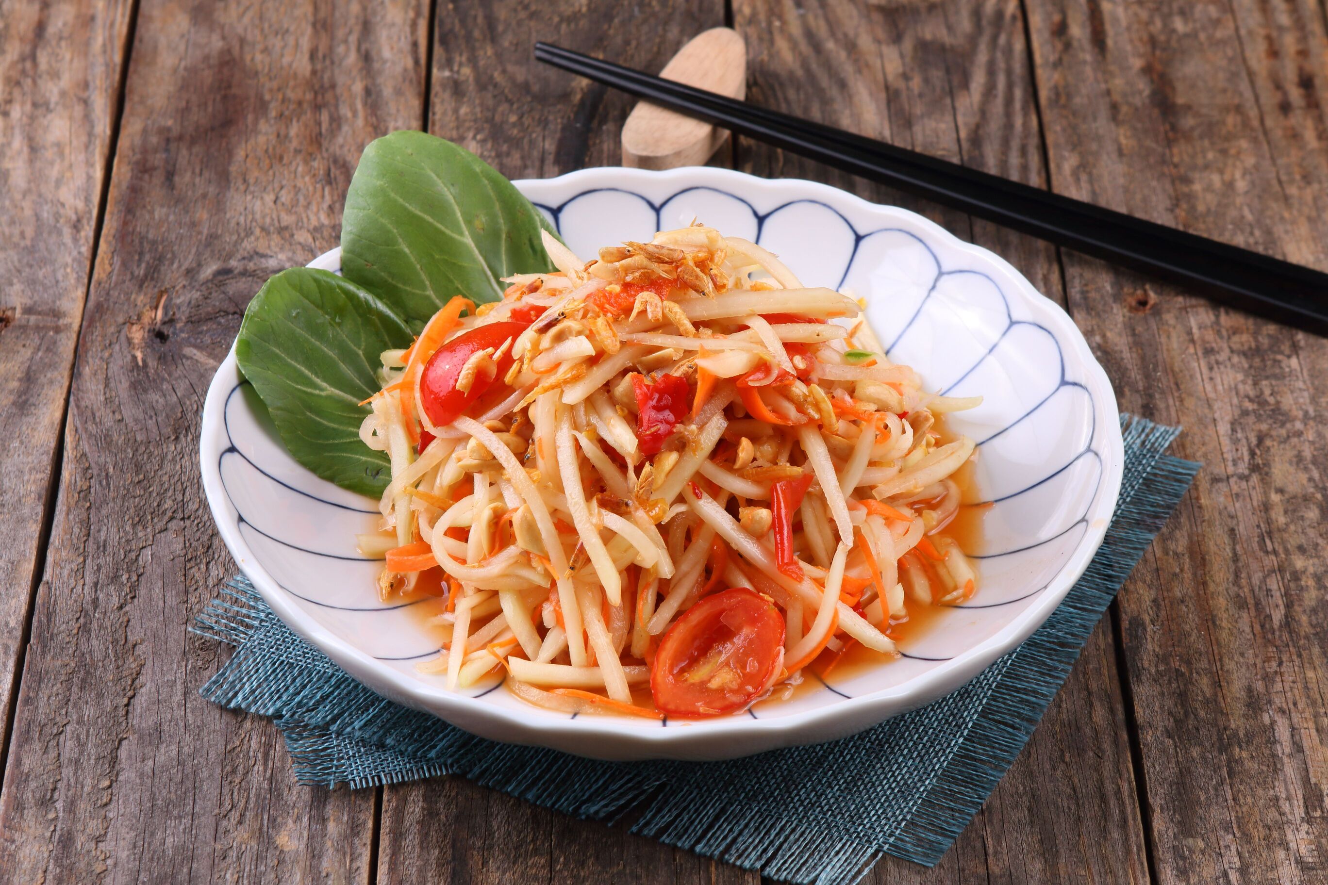 Close-up of Thai green papaya salad with tomato wedges in a white bowl with blue patterns, chopsticks beside it on a dark wooden table